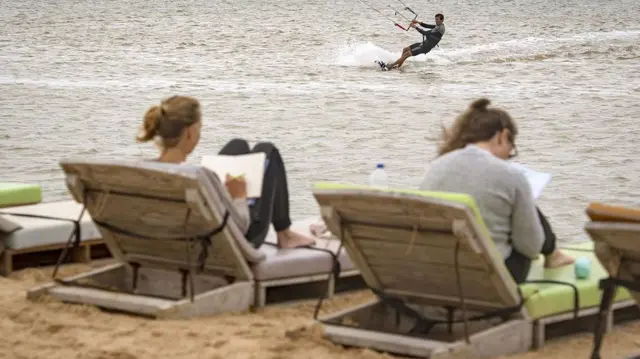 Deux touristes profitent des paysages du Sahara occidental : l'un se détend sur une chaise longue en bord de mer tandis que l'autre pratique le kitesurf dans l'océan Atlantique